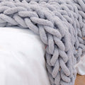 The Anchor chunky knit blanket in grey, draped over the corner of a white bed, showing the thick braided weave up close